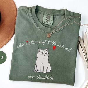 Pode incluir: Uma camiseta verde com uma ilustra&ccedil;&atilde;o de um gato branco e o texto "who's afraid of little old me? you should be".