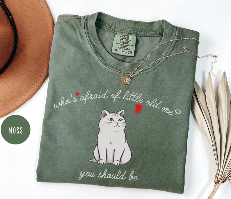 Pode incluir: Uma camiseta verde com uma ilustra&ccedil;&atilde;o de um gato branco e o texto "who's afraid of little old me? you should be".