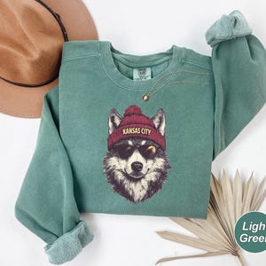 May include: A light green sweatshirt with a wolf wearing sunglasses and a red beanie that says "Kansas City".