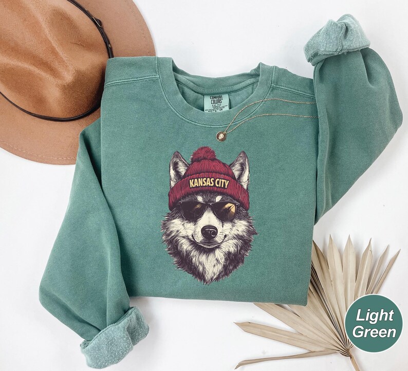 May include: A light green sweatshirt with a wolf wearing sunglasses and a red beanie that says "Kansas City".