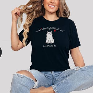 Pode incluir: Uma camiseta preta com uma ilustra&ccedil;&atilde;o de um gato branco e o texto "who's afraid of little old me? you should be".