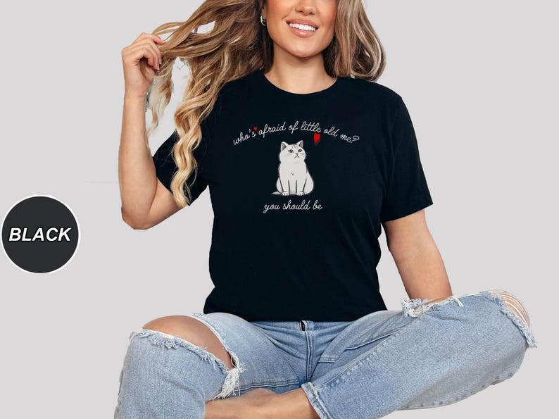 Pode incluir: Uma camiseta preta com uma ilustra&ccedil;&atilde;o de um gato branco e o texto "who's afraid of little old me? you should be".