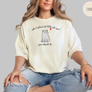 Pode incluir: Uma mulher vestindo uma camiseta branca com uma ilustra&ccedil;&atilde;o de um gato e o texto "who's afraid of little old me? you should be". A camiseta &eacute; de cor marfim.