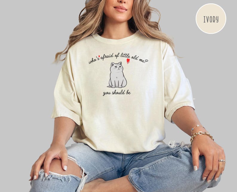 Pode incluir: Uma mulher vestindo uma camiseta branca com uma ilustra&ccedil;&atilde;o de um gato e o texto "who's afraid of little old me? you should be". A camiseta &eacute; de cor marfim.