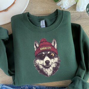 May include: A forest green sweatshirt with a graphic of a wolf wearing a red beanie with the text "Kansas City" on it. The wolf is wearing sunglasses.