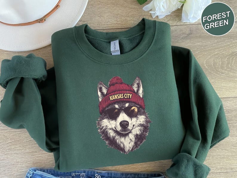 May include: A forest green sweatshirt with a graphic of a wolf wearing a red beanie with the text "Kansas City" on it. The wolf is wearing sunglasses.