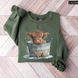 Puede incluir: Una sudadera verde con un gr&aacute;fico de una vaca Highland tomando un ba&ntilde;o en un cubo de metal. La vaca es marr&oacute;n y blanca con una expresi&oacute;n linda.