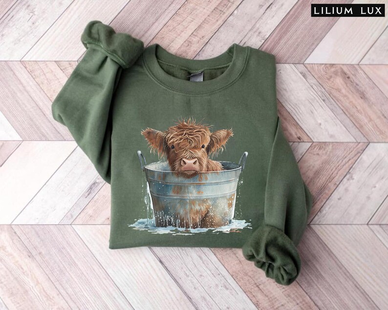 Puede incluir: Una sudadera verde con un gr&aacute;fico de una vaca Highland tomando un ba&ntilde;o en un cubo de metal. La vaca es marr&oacute;n y blanca con una expresi&oacute;n linda.