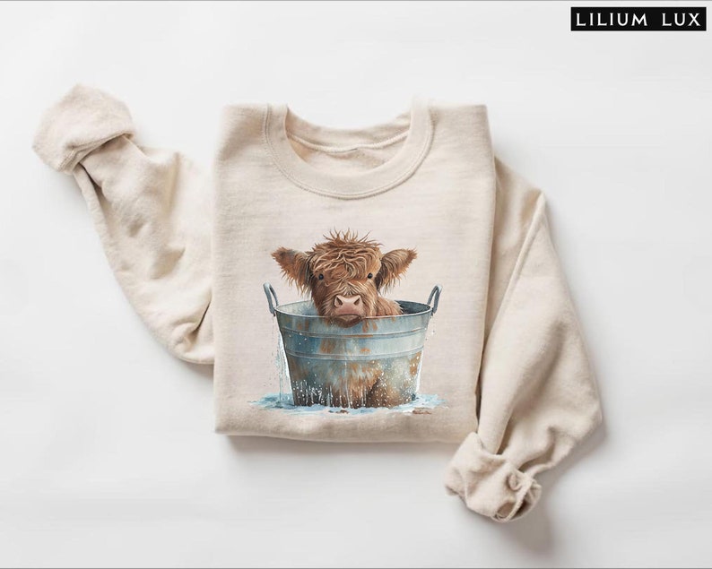 Puede incluir: Una sudadera beige con un gr&aacute;fico de una vaca Highland marr&oacute;n y blanca tomando un ba&ntilde;o en un cubo de metal. La vaca est&aacute; mirando a la c&aacute;mara con una expresi&oacute;n curiosa.