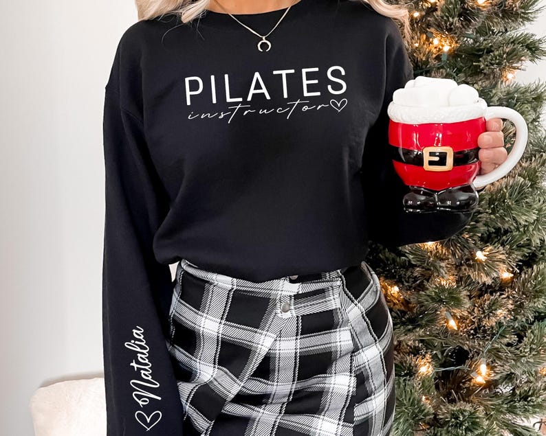 May include: Black sweatshirt with the text "PILATES instructor" in white lettering and a small heart. The sweatshirt has long sleeves with the name "Natalia" written on the left sleeve. The person is wearing a black and white plaid skirt.