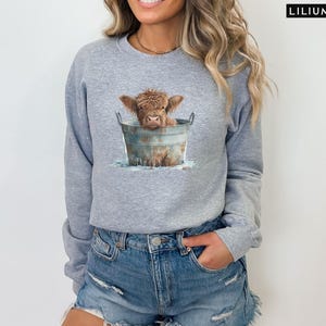 Puede incluir: Una sudadera gris con un gr&aacute;fico de una vaca Highland marr&oacute;n en un cubo de metal. La vaca est&aacute; mirando a la c&aacute;mara y tiene la nariz mojada y marr&oacute;n.