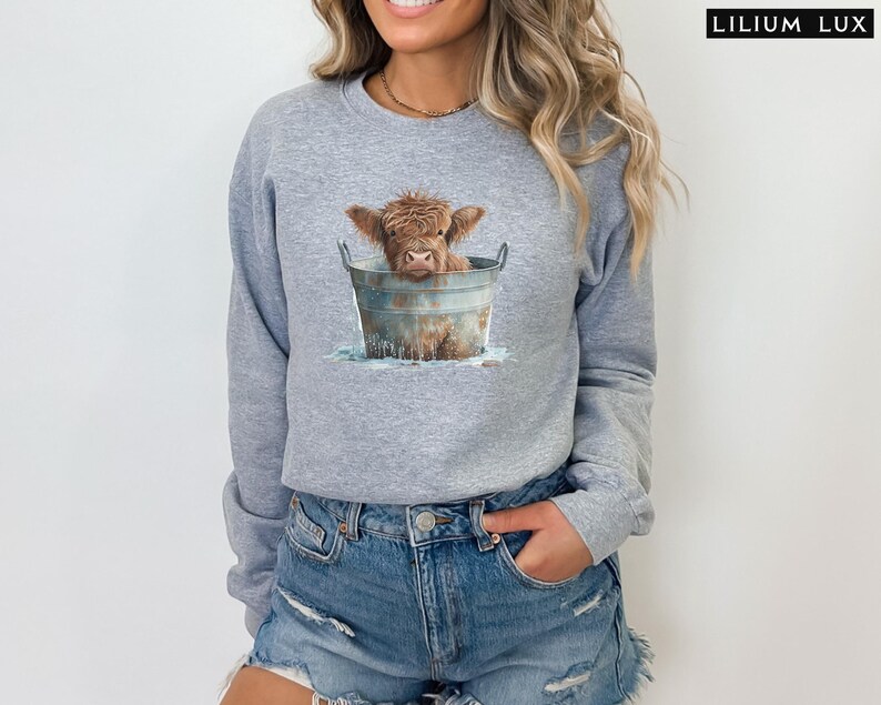 Puede incluir: Una sudadera gris con un gr&aacute;fico de una vaca Highland marr&oacute;n en un cubo de metal. La vaca est&aacute; mirando a la c&aacute;mara y tiene la nariz mojada y marr&oacute;n.