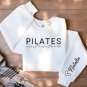 May include: White sweatshirt with the text "PILATES instructor" and a heart on the front. The left sleeve has a heart and the text "Natalia" on it.