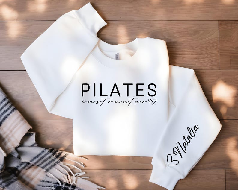 May include: White sweatshirt with the text "PILATES instructor" and a heart on the front. The left sleeve has a heart and the text "Natalia" on it.