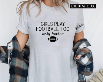May include: A light gray t-shirt with the text "GIRLS PLAY FOOTBALL TOO -only better-" and a football graphic. The shirt is a crew neck style and is being worn by a person. The brand name "LILIUM LUX" is in the top right corner.