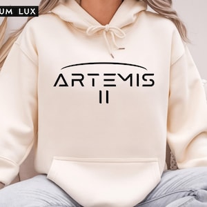 May include: Cream-colored hoodie with a drawstring hood and a front pocket. The black text "ARTEMIS II" is printed across the chest. The brand name "LILIUM LUX" is in the upper left corner.