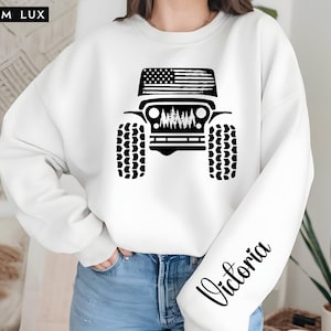 May include: White sweatshirt featuring a black graphic of a Jeep with an American flag on top. The Jeep design includes tires and a forest scene. The name "Victoria" is printed in black script on the sleeve. The brand name "LILIUM LUX" is in the top left corner.