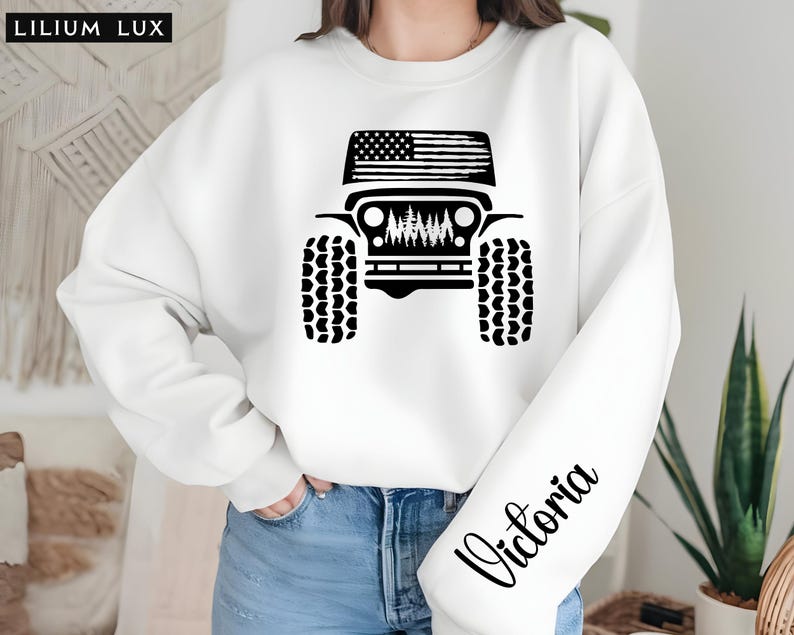 May include: White sweatshirt featuring a black graphic of a Jeep with an American flag on top. The Jeep design includes tires and a forest scene. The name "Victoria" is printed in black script on the sleeve. The brand name "LILIUM LUX" is in the top left corner.