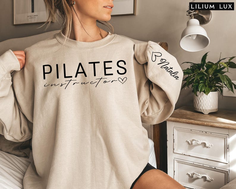 May include: A beige sweatshirt with the text "PILATES instructor" and a heart on the front. The sleeve has the text "Natalia" with a heart.