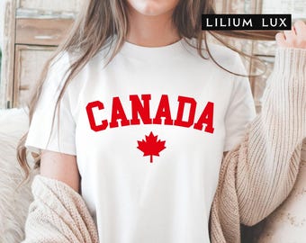 May include: White t-shirt with the word "CANADA" in red block letters and a red maple leaf graphic. The shirt is short-sleeved and appears to be made of cotton. The brand name "LILIUM LUX" is in the top right corner.