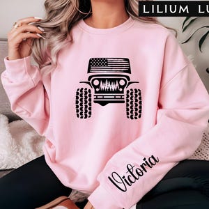 May include: A light pink sweatshirt featuring a black graphic of a Jeep with an American flag on the roof and a forest scene. The word "Victoria" is written in cursive on the sleeve. The text "LILIUM LUX" is at the top.
