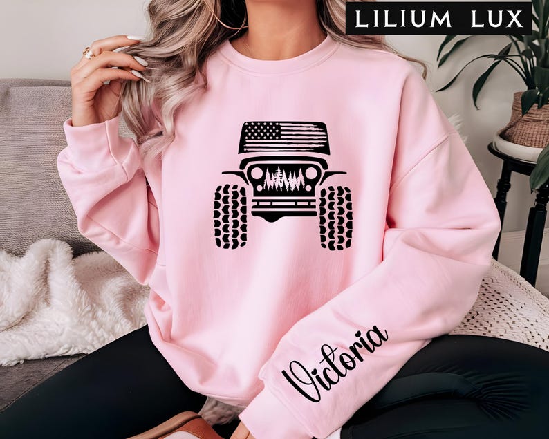 May include: A light pink sweatshirt featuring a black graphic of a Jeep with an American flag on the roof and a forest scene. The word "Victoria" is written in cursive on the sleeve. The text "LILIUM LUX" is at the top.
