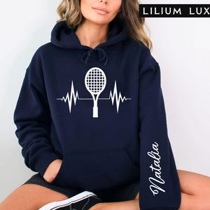 May include: Navy blue hoodie with a white tennis racket and heartbeat graphic. The sleeve features the name "Natalia" in a cursive font. The text "LILIUM LUX" is at the top right.