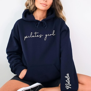 May include: A navy blue hoodie with the text "pilates girl" in white script on the front and the name "Natalia" in white script on the left sleeve.