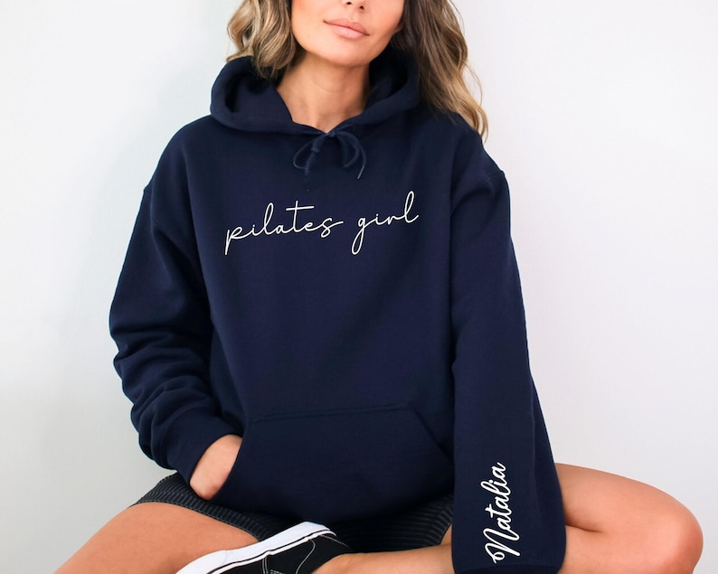 May include: A navy blue hoodie with the text "pilates girl" in white script on the front and the name "Natalia" in white script on the left sleeve.