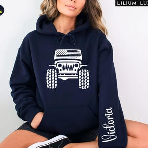 May include: Navy blue hoodie with a white graphic of a Jeep featuring an American flag design on the top and a forest scene. The sleeve has the name "Victoria" in a script font. The hoodie has a front pocket and a hood.