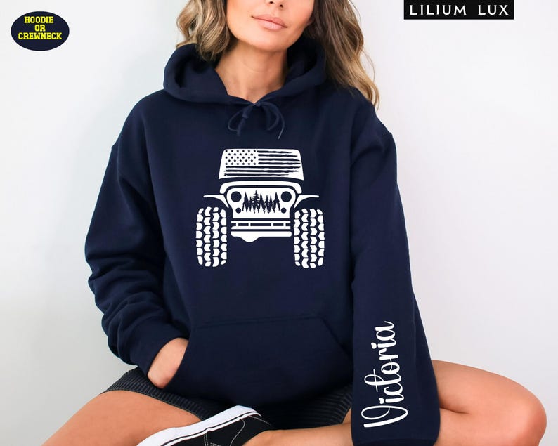 May include: Navy blue hoodie with a white graphic of a Jeep featuring an American flag design on the top and a forest scene. The sleeve has the name "Victoria" in a script font. The hoodie has a front pocket and a hood.