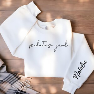 May include: White sweatshirt with the text "pilates girl" in black script on the front and the name "Natalia" in black script on the sleeve.
