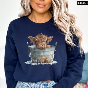 Puede incluir: Una sudadera azul marino con un gr&aacute;fico de una vaca Highland tomando un ba&ntilde;o en un cubo. La vaca es marr&oacute;n y blanca y tiene una expresi&oacute;n linda en su rostro.