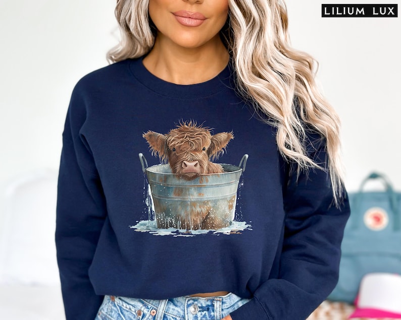Puede incluir: Una sudadera azul marino con un gr&aacute;fico de una vaca Highland tomando un ba&ntilde;o en un cubo. La vaca es marr&oacute;n y blanca y tiene una expresi&oacute;n linda en su rostro.