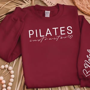 May include: Burgundy red sweatshirt with white text that reads "PILATES instructor" with a heart symbol. The right sleeve has white text that reads "B Natalia" with a heart symbol.
