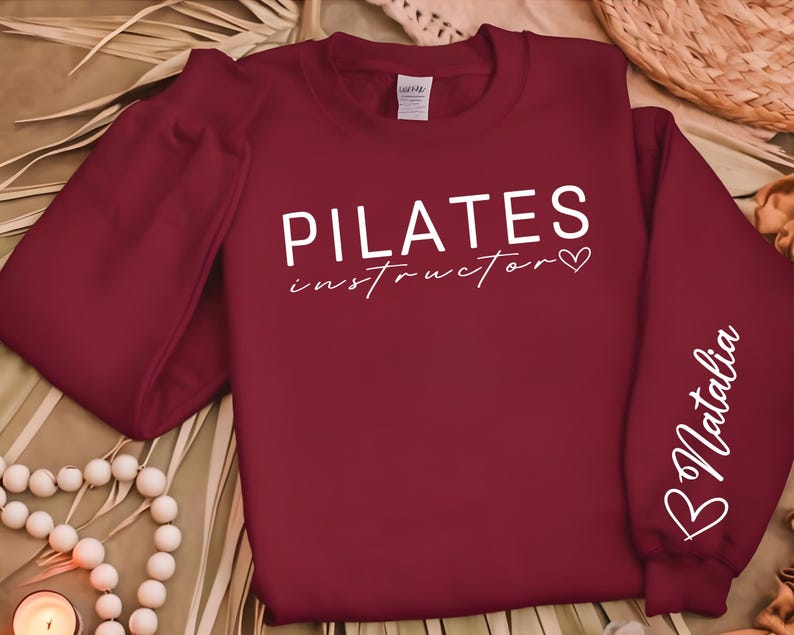 May include: Burgundy red sweatshirt with white text that reads "PILATES instructor" with a heart symbol. The right sleeve has white text that reads "B Natalia" with a heart symbol.