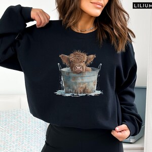 Puede incluir: Una sudadera negra con un gr&aacute;fico de una vaca Highland marr&oacute;n y blanca en un cubo de metal. La vaca est&aacute; mirando a la c&aacute;mara y tiene la cabeza inclinada hacia un lado. El cubo est&aacute; lleno de agua y hay salpicaduras de agua alrededor de la vaca.