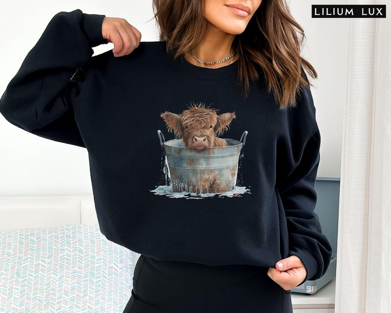 Puede incluir: Una sudadera negra con un gr&aacute;fico de una vaca Highland marr&oacute;n y blanca en un cubo de metal. La vaca est&aacute; mirando a la c&aacute;mara y tiene la cabeza inclinada hacia un lado. El cubo est&aacute; lleno de agua y hay salpicaduras de agua alrededor de la vaca.