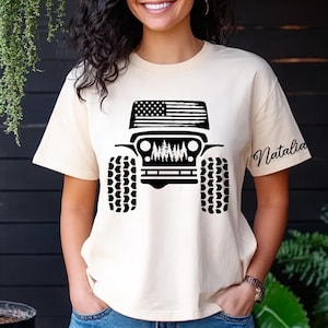 May include: A white t-shirt with a black silhouette of a Jeep with an American flag on the hood and trees in the grill. The text "Natalia" is on the sleeve of the shirt.