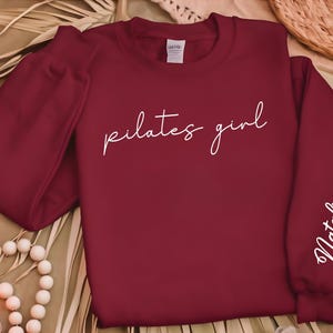 May include: Burgundy sweatshirt with the text "pilates girl" in white script on the front and the name "Natalia" in white script on the right sleeve.