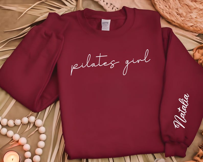 May include: Burgundy sweatshirt with the text "pilates girl" in white script on the front and the name "Natalia" in white script on the right sleeve.