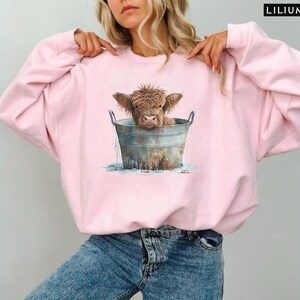Puede incluir: Una sudadera rosa con un gr&aacute;fico de una vaca Highland marr&oacute;n y blanca en un cubo de metal. La vaca est&aacute; mirando a la c&aacute;mara y tiene la cabeza y los hombros en el cubo. El cubo est&aacute; lleno de agua y hay salpicaduras de agua alrededor de la vaca.