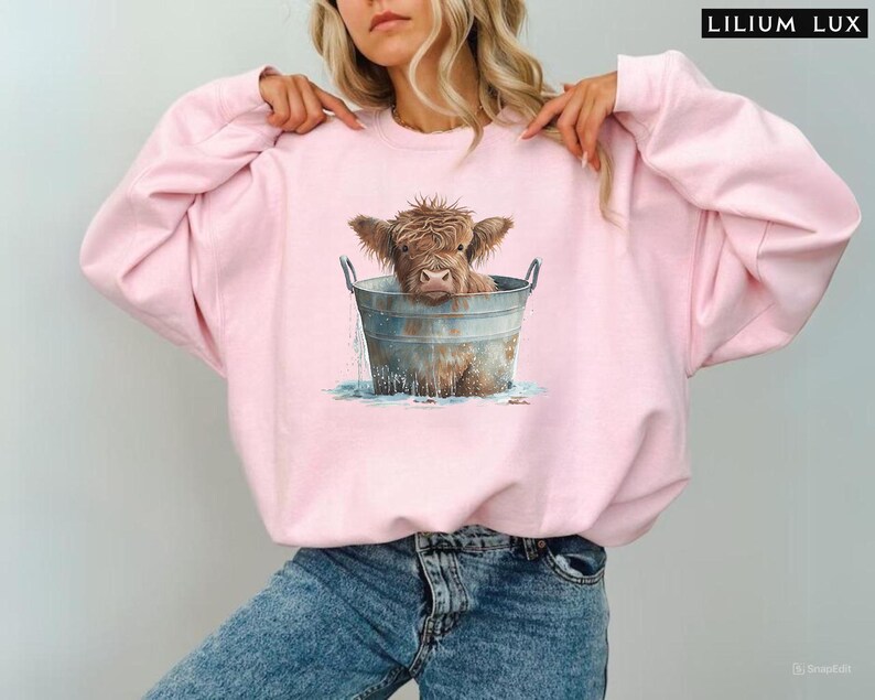 Puede incluir: Una sudadera rosa con un gr&aacute;fico de una vaca Highland marr&oacute;n y blanca en un cubo de metal. La vaca est&aacute; mirando a la c&aacute;mara y tiene la cabeza y los hombros en el cubo. El cubo est&aacute; lleno de agua y hay salpicaduras de agua alrededor de la vaca.