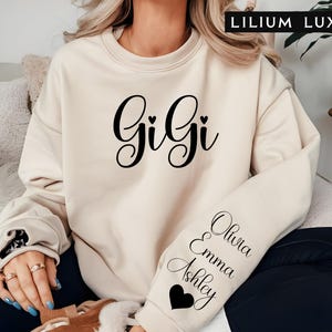 May include: A cream-colored sweatshirt with the word "Gigi" in black cursive lettering and two small hearts. The left sleeve has the names "Olivia", "Emma", and "Ashley" written in black cursive lettering with a heart at the bottom.