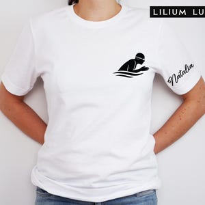 May include: White t-shirt featuring a black silhouette of a swimmer in the water. The text "LILIUM LUX" is at the top right, and the name "Natalia" is written on the right sleeve in cursive.