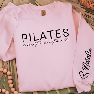 May include: Pink sweatshirt with the text "PILATES instructor" and a small heart on the front. The left sleeve has the name "Natalia" written on it.