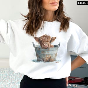 Puede incluir: Sudadera blanca con un gr&aacute;fico de una vaca Highland marr&oacute;n y blanca en un cubo de metal. La vaca est&aacute; mirando a la c&aacute;mara y tiene la cabeza y los hombros en el cubo. El cubo est&aacute; lleno de agua y la vaca est&aacute; rodeada de gotas de agua.