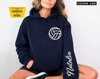Op de afbeelding: Een marineblauwe hoodie met een witte volleybalafbeelding op de borst en de naam Natalia in wit schrift op de linkermouw. De tekst LILIUM LUX staat rechtsboven en een geel kader geeft informatie over de beschikbaarheid van het item.