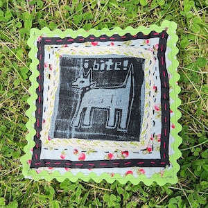 May include: A green, black, and white fabric square with a stitched border. The center features a blue denim patch with a black and white illustration of a dog and the text "I bite!"