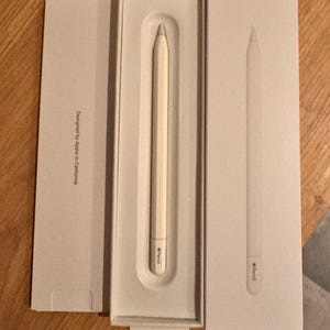 Op de afbeelding: Een gebroken witte Apple Pencil in de originele verpakking. De pen wordt weergegeven in een open doos met de tekst "Designed by Apple in California" zichtbaar op de verpakking. De pen is lichtcrème van kleur met een zilveren punt.